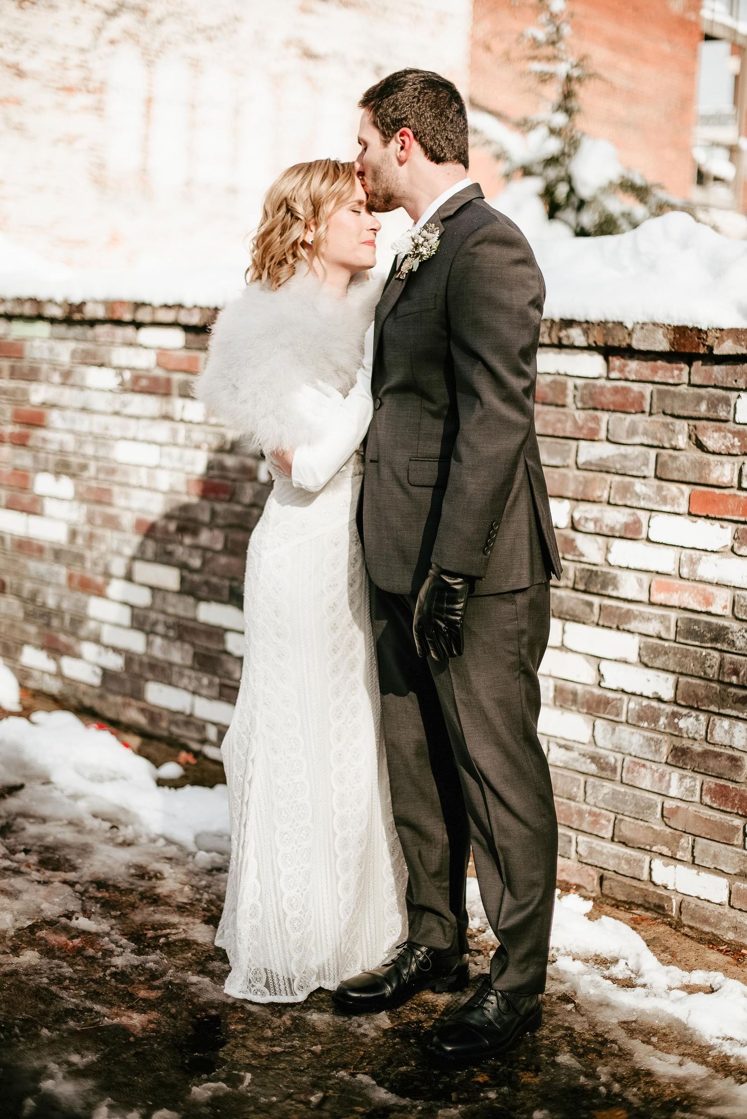 Bride and groom share a romantic winter kiss in snow, wearing elegant wedding attire beside a rustic brick wall.