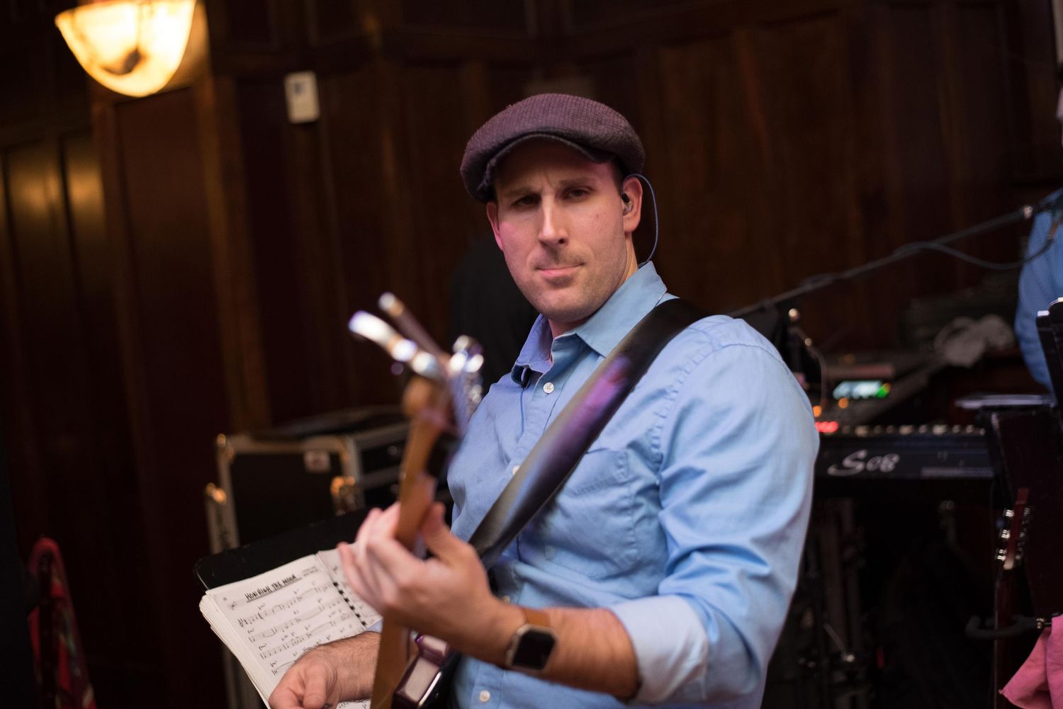 Musician playing electric guitar while reading sheet music, wearing a blue shirt and flat cap at an indoor venue.