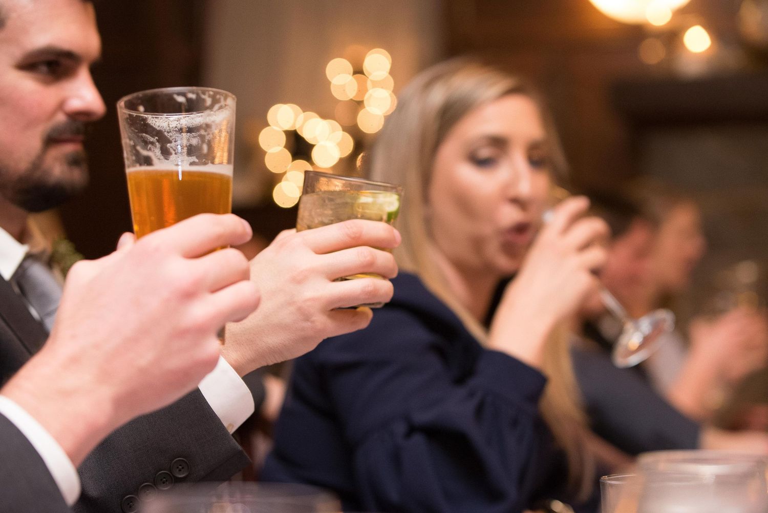 People toasting with beer and cocktails at a festive indoor gathering with warm bokeh lights in the background.