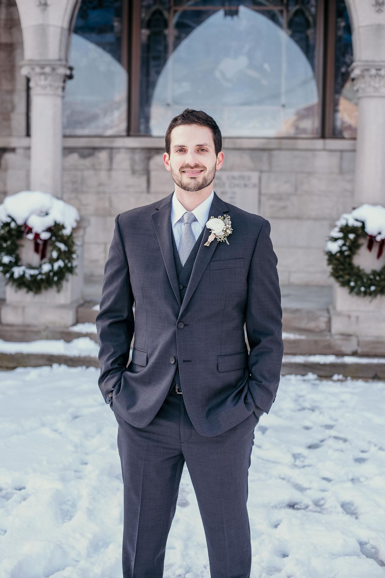 Groom in charcoal suit with boutonniere poses in snow outside stone church with Christmas wreaths.