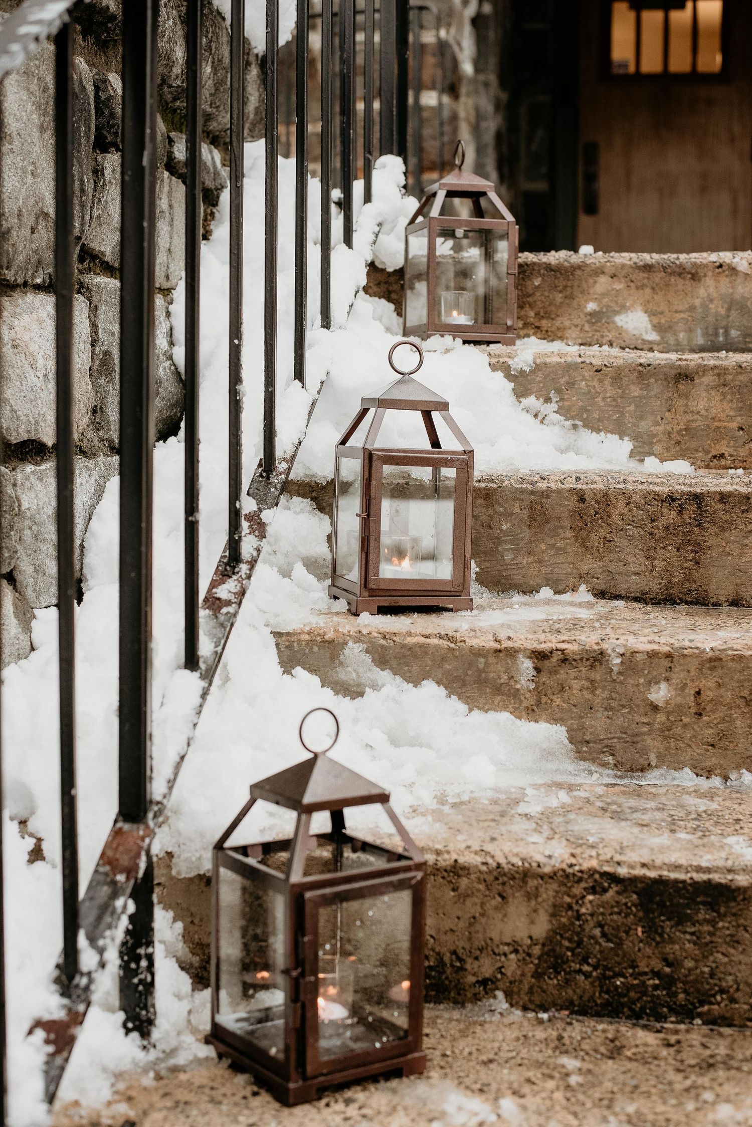 Rustic bronze lanterns with candles line snow-covered stone steps, creating a warm winter ambiance.