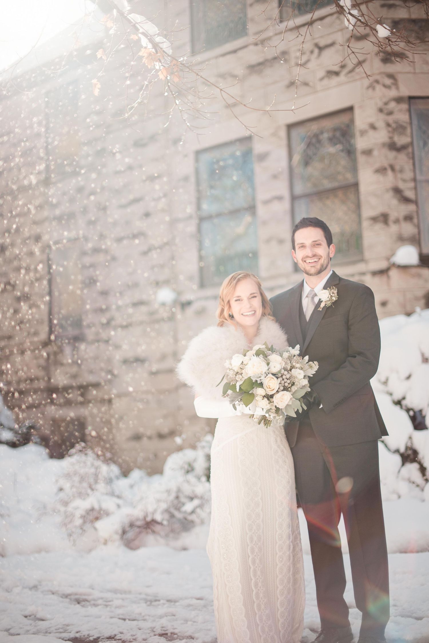 Couple posing in snow for winter wedding, bride holding white bouquet wearing fur wrap, groom in dark suit beside stone building.
