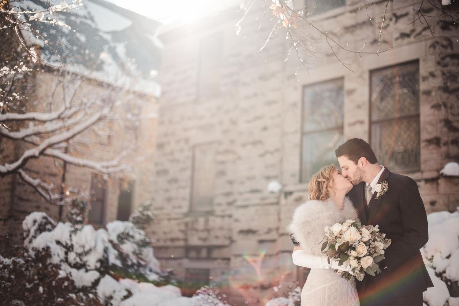Bride and groom share a romantic winter kiss in the snow outside a stone building on their wedding day.