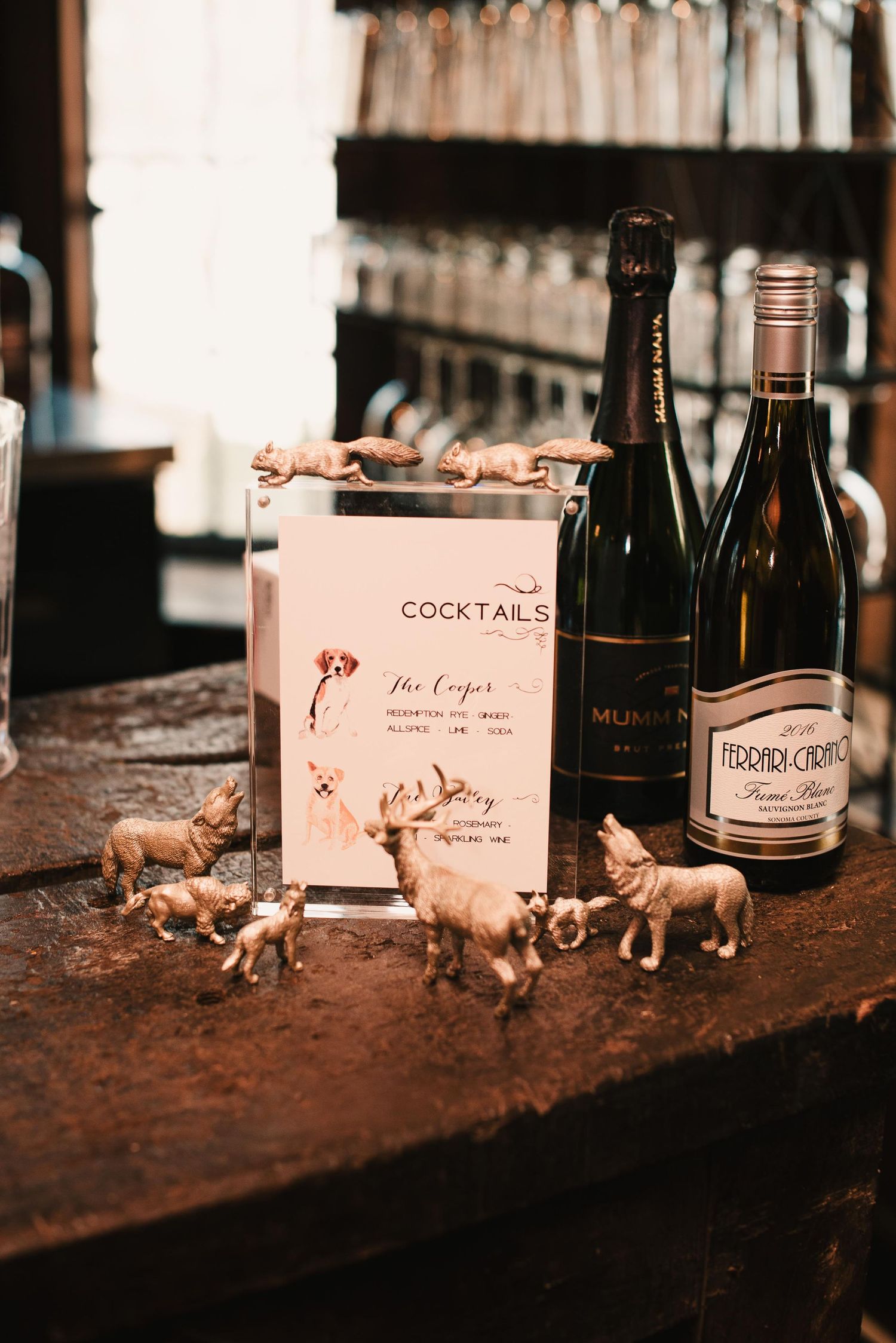 Cocktail menu display with gold animal figurines, Mumm and Ferrari-Carano wine bottles on rustic bar.