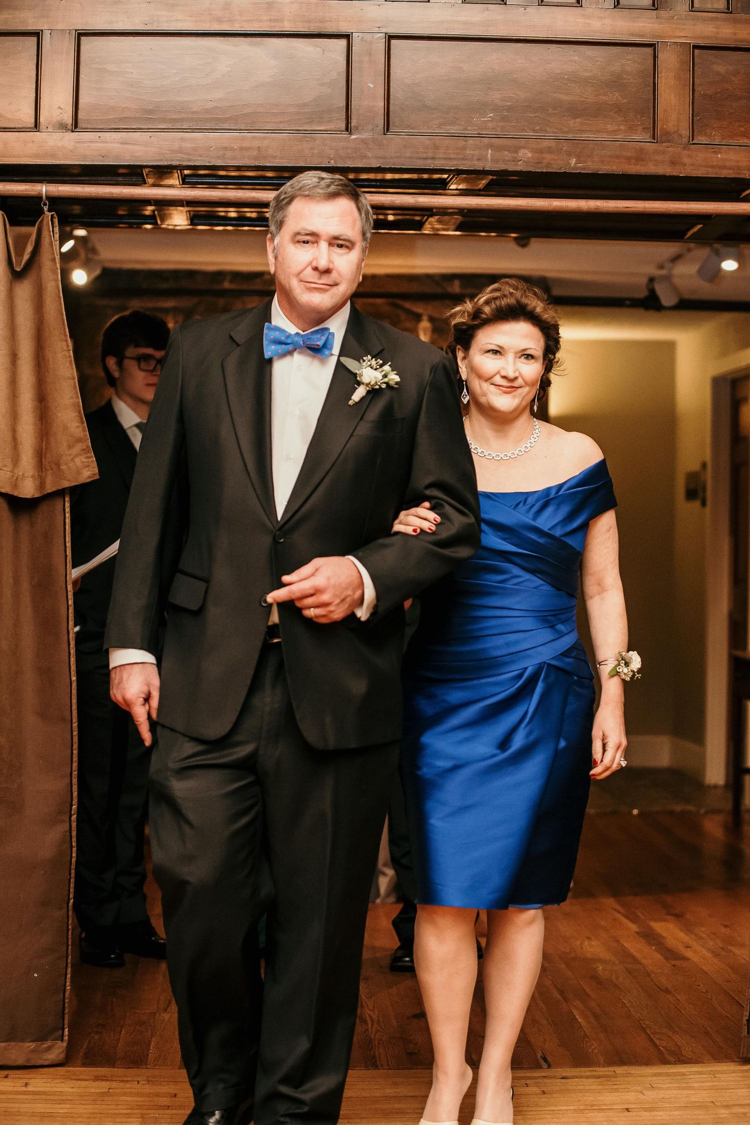 Elegant couple at wedding reception, man in black tuxedo with blue bow tie, woman in royal blue off-shoulder dress.