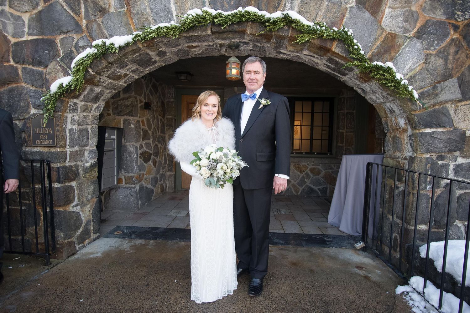 Bride and groom posing under stone arch at winter wedding venue, bride holding white bouquet.