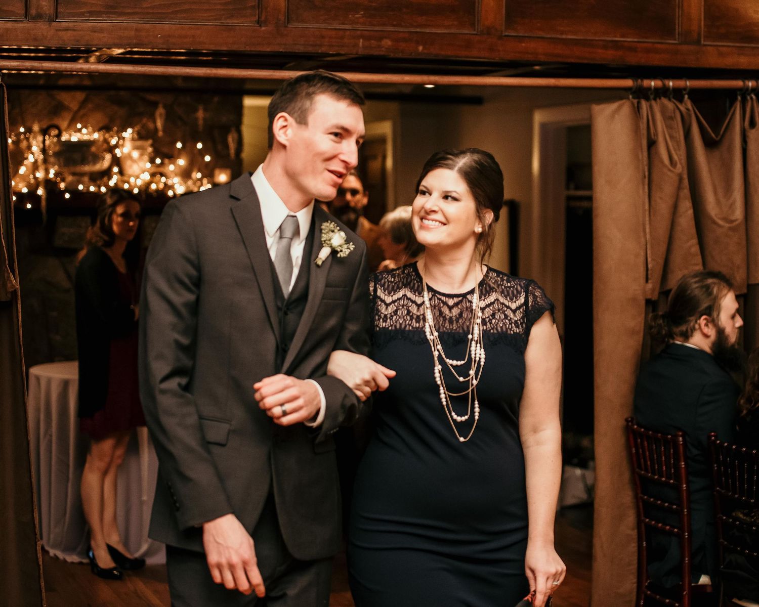 Couple in formal attire smiling at a wedding reception, woman in black dress with pearls, man in gray suit.