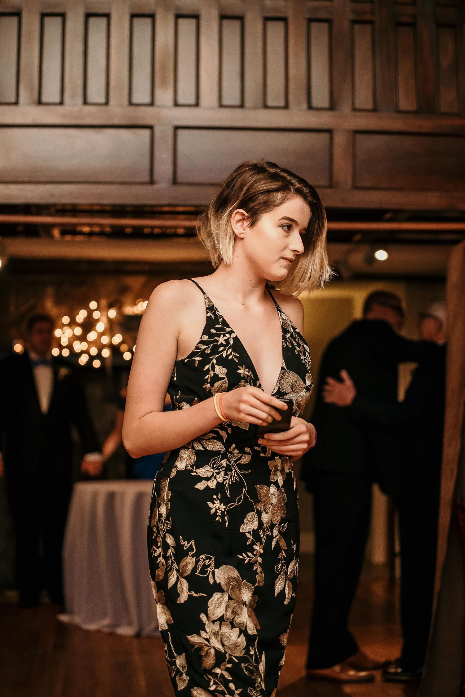 A young woman in a black floral spaghetti strap dress stands elegantly at an upscale indoor evening event.