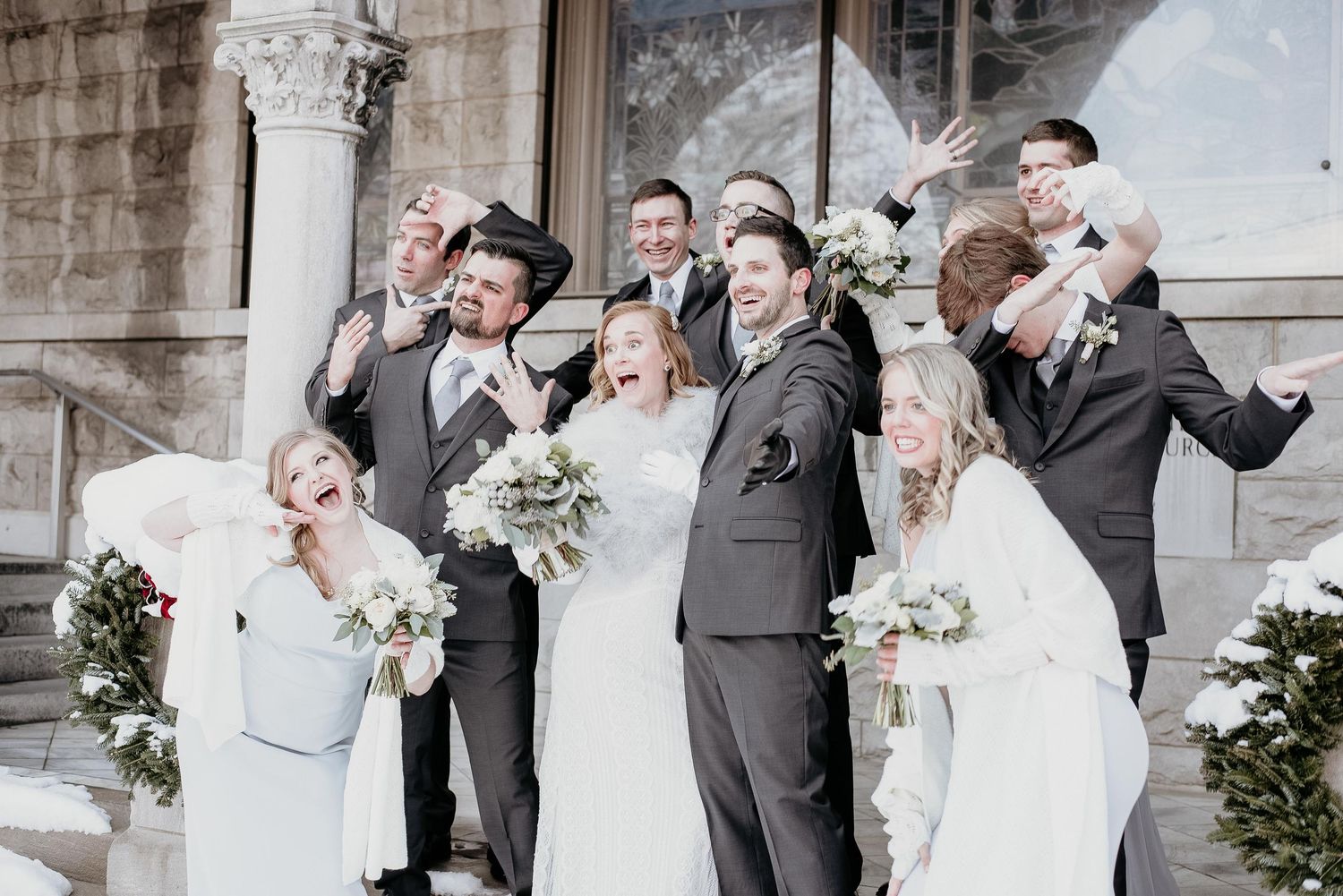 Wedding party celebrating joyfully on church steps, bride holding white bouquet, groomsmen in gray suits, bridesmaids in white dresses.