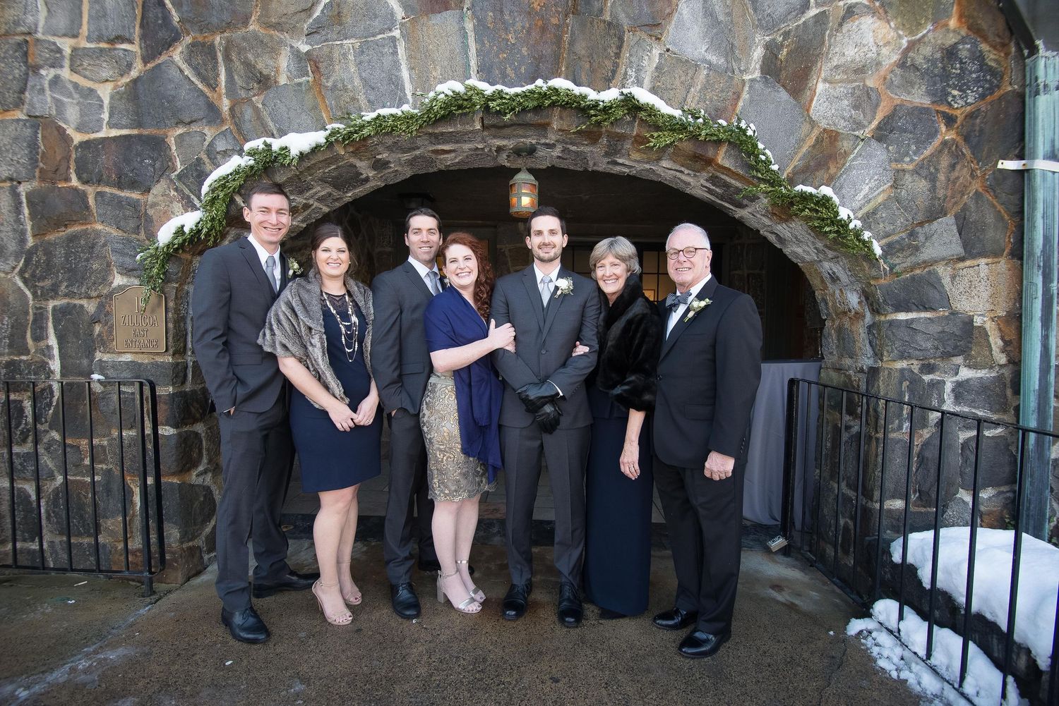 Group of seven formally dressed people posing together under a stone archway decorated with winter greenery.