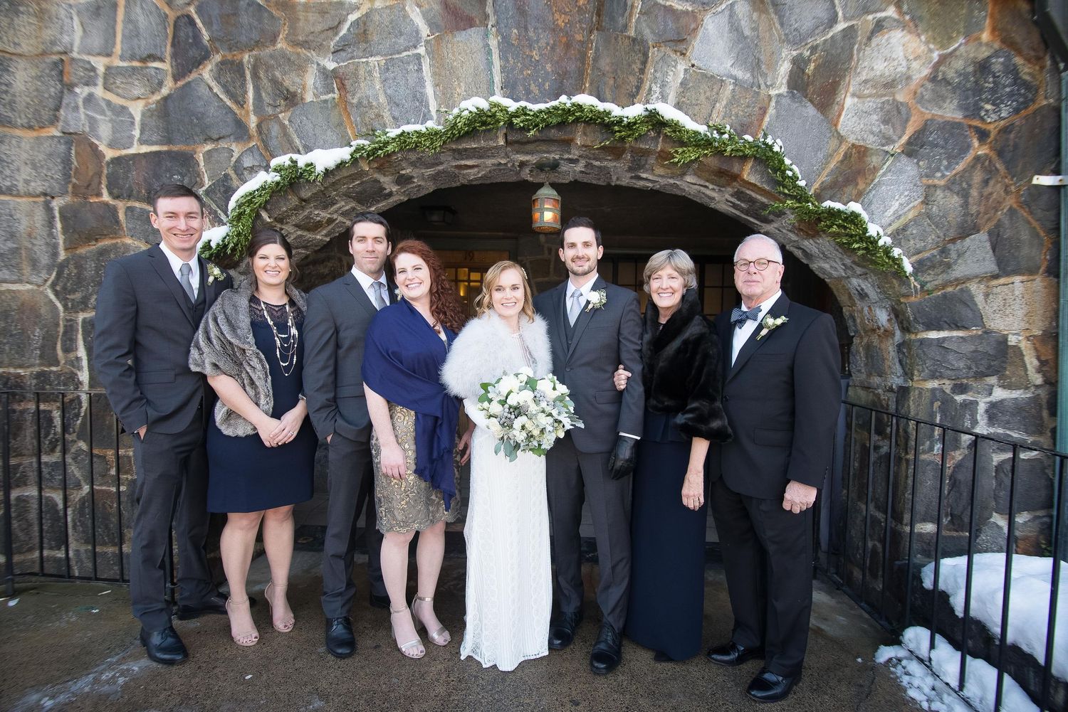 Wedding party poses in front of a stone arch entrance in winter, bride holding white bouquet with family members.