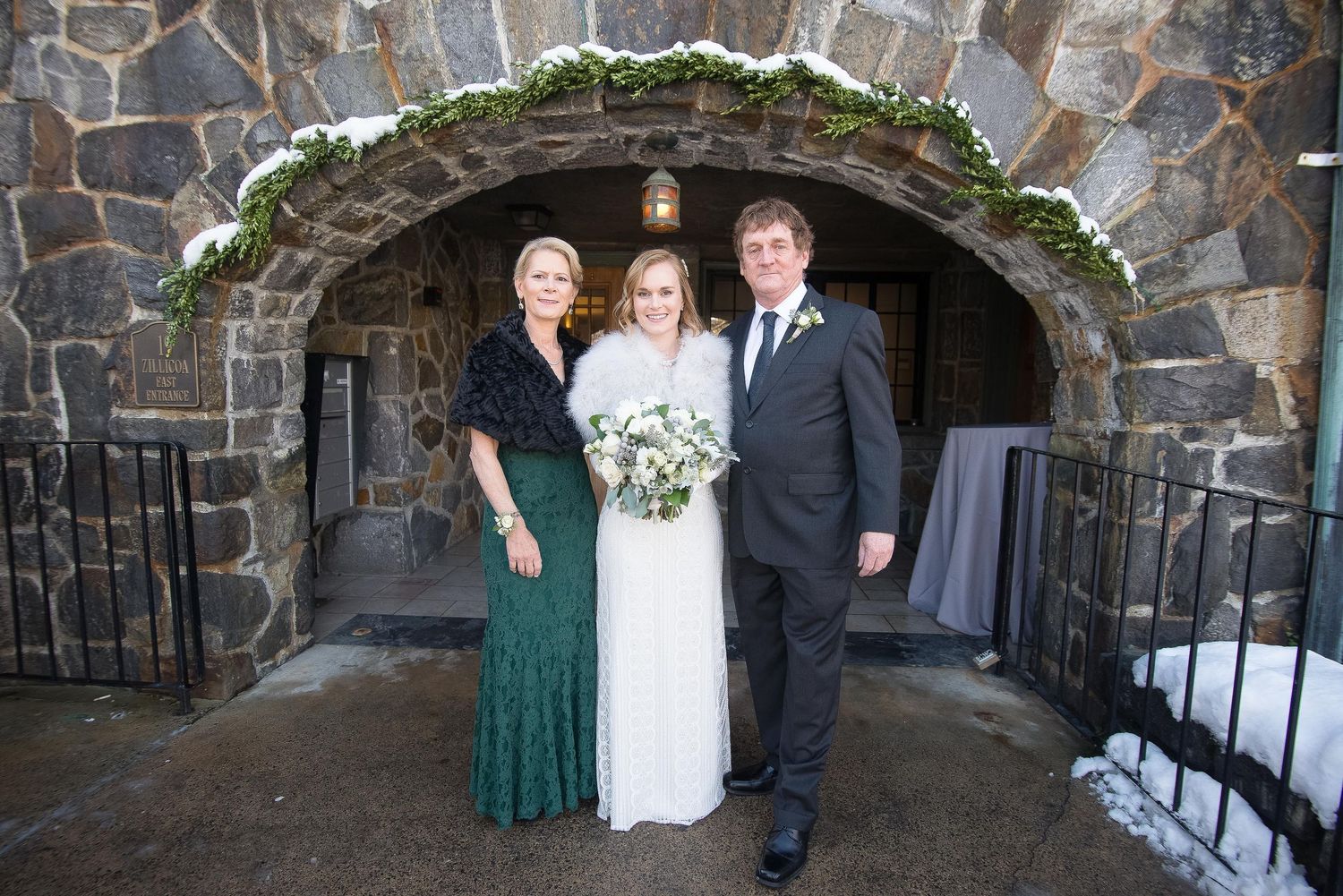 Bride and groom pose with a guest in front of a stone arch doorway decorated with greenery at a winter wedding.