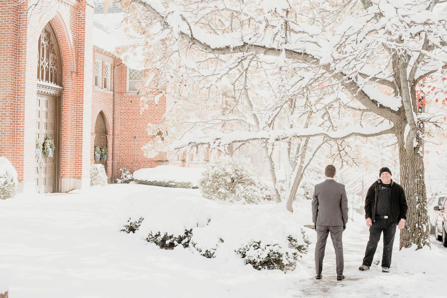 Two men stand outside a snow-covered brick church surrounded by snow-laden trees in a winter wonderland setting.