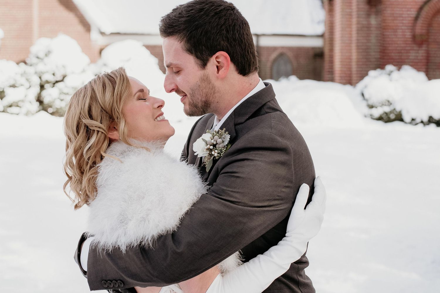 A couple embraces in a snowy winter wedding scene, the bride in a white fur wrap and groom in a gray suit with boutonniere.