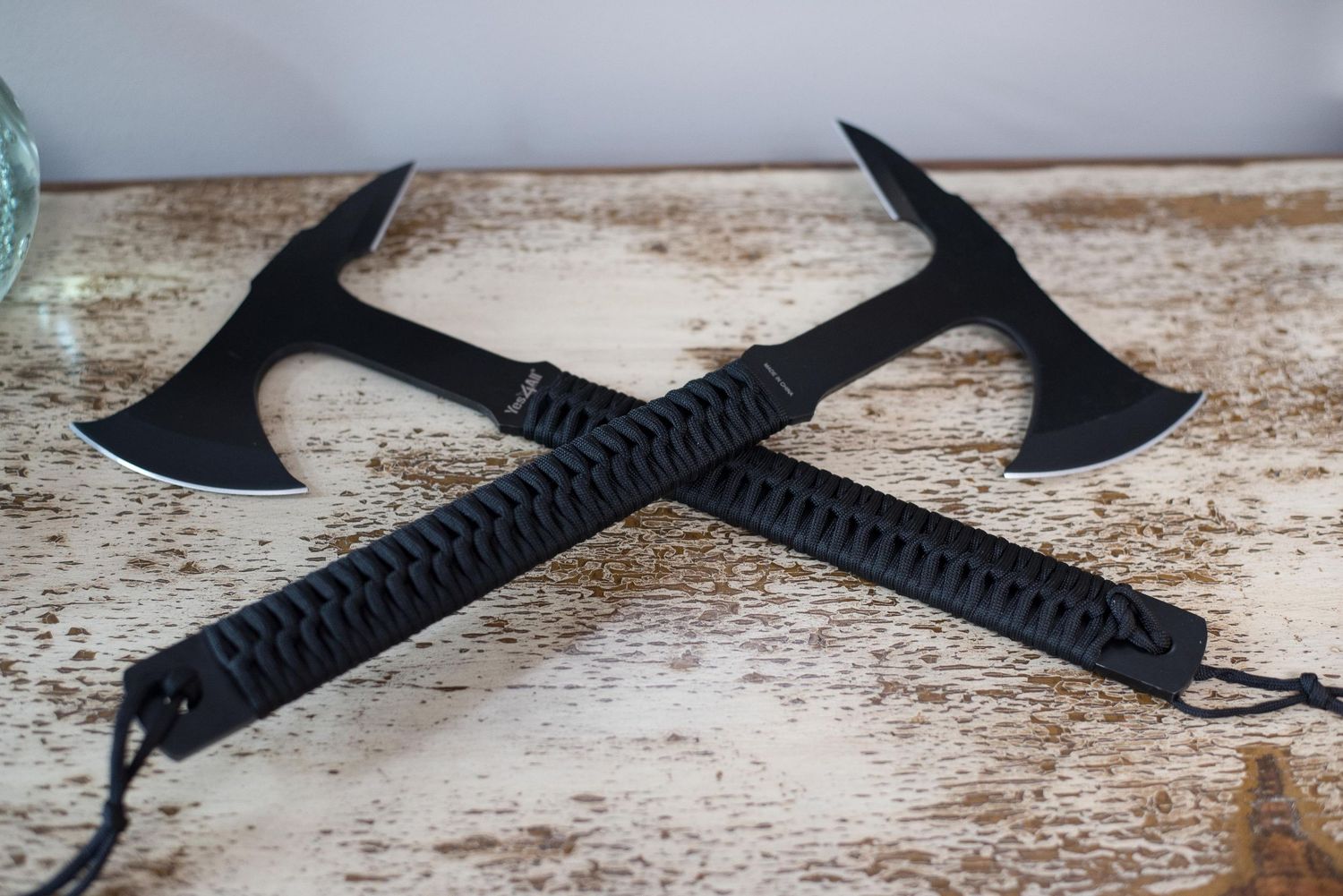 Two black tactical tomahawk axes with paracord wrapped handles crossed on a rustic wooden surface.