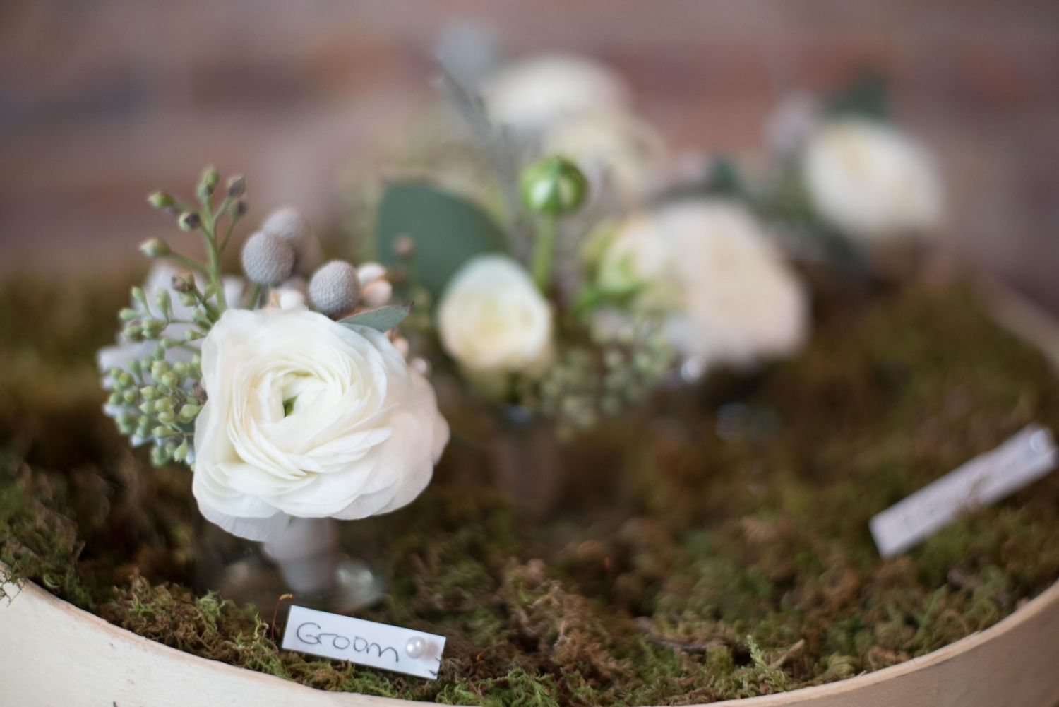 White ranunculus boutonnières on a moss-covered wooden tray, perfect for rustic wedding floral arrangements.