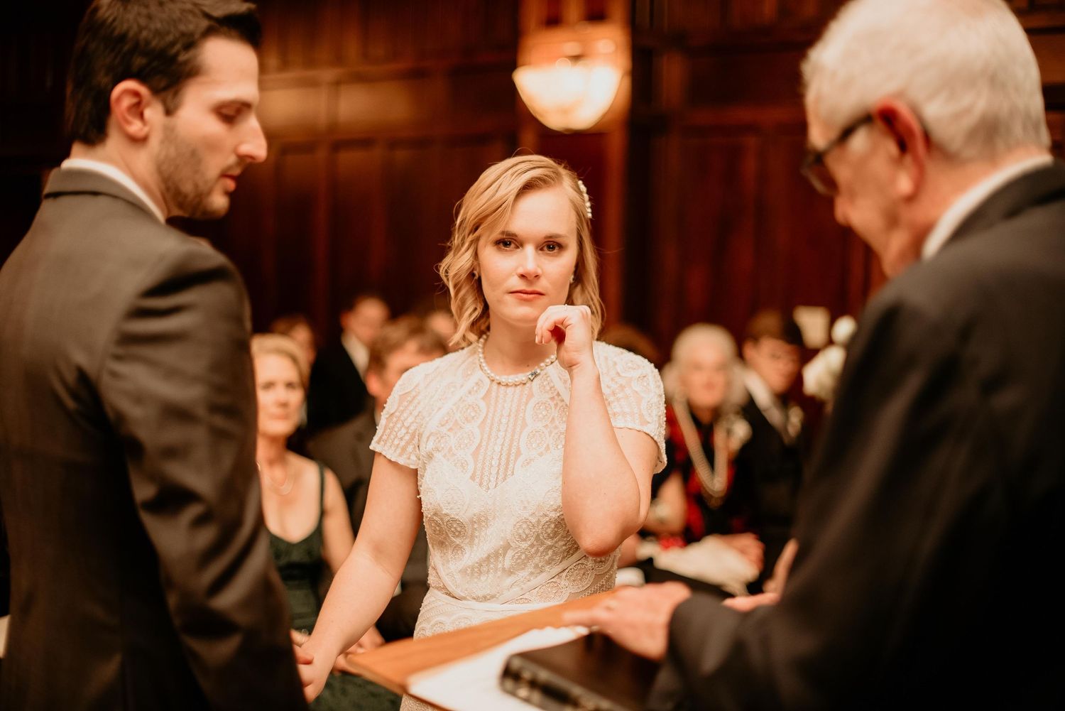 Bride in beaded gown listens attentively during wedding ceremony while groom and officiant stand nearby in elegant venue.