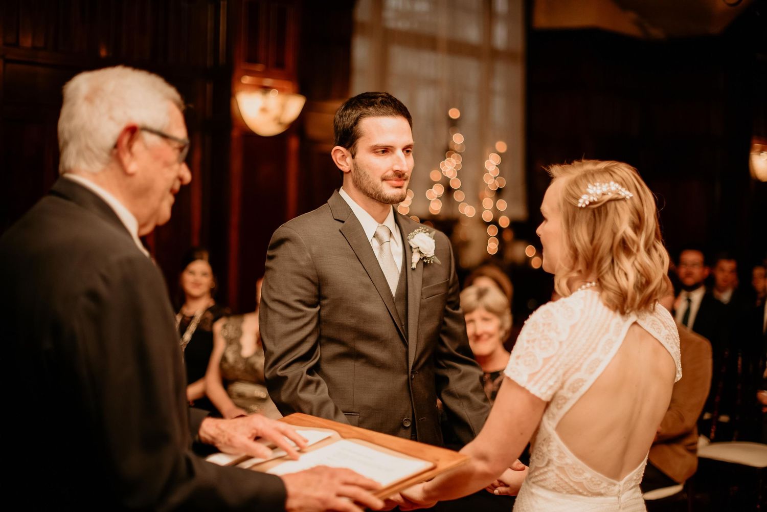 Groom smiling at bride during indoor wedding ceremony with officiant and guests in warm-lit venue.