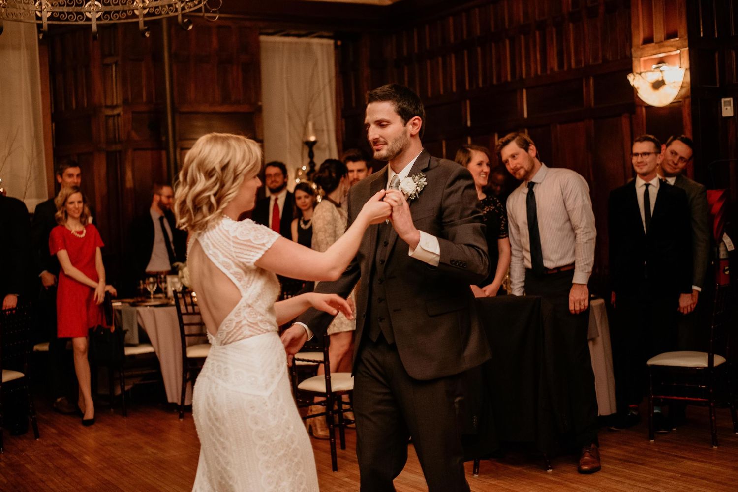 Bride and groom share their first dance at elegant wedding reception while guests watch in warmly lit ballroom.