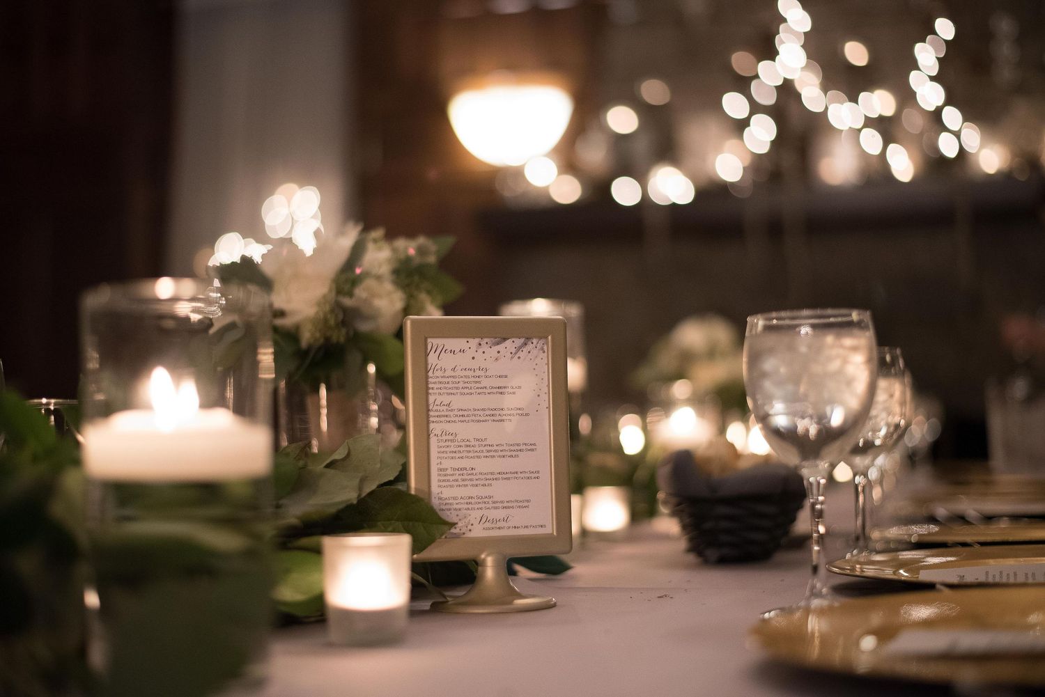 Elegant wedding reception table with glowing candles, greenery, wine glasses, and a framed menu card in warm bokeh lighting.