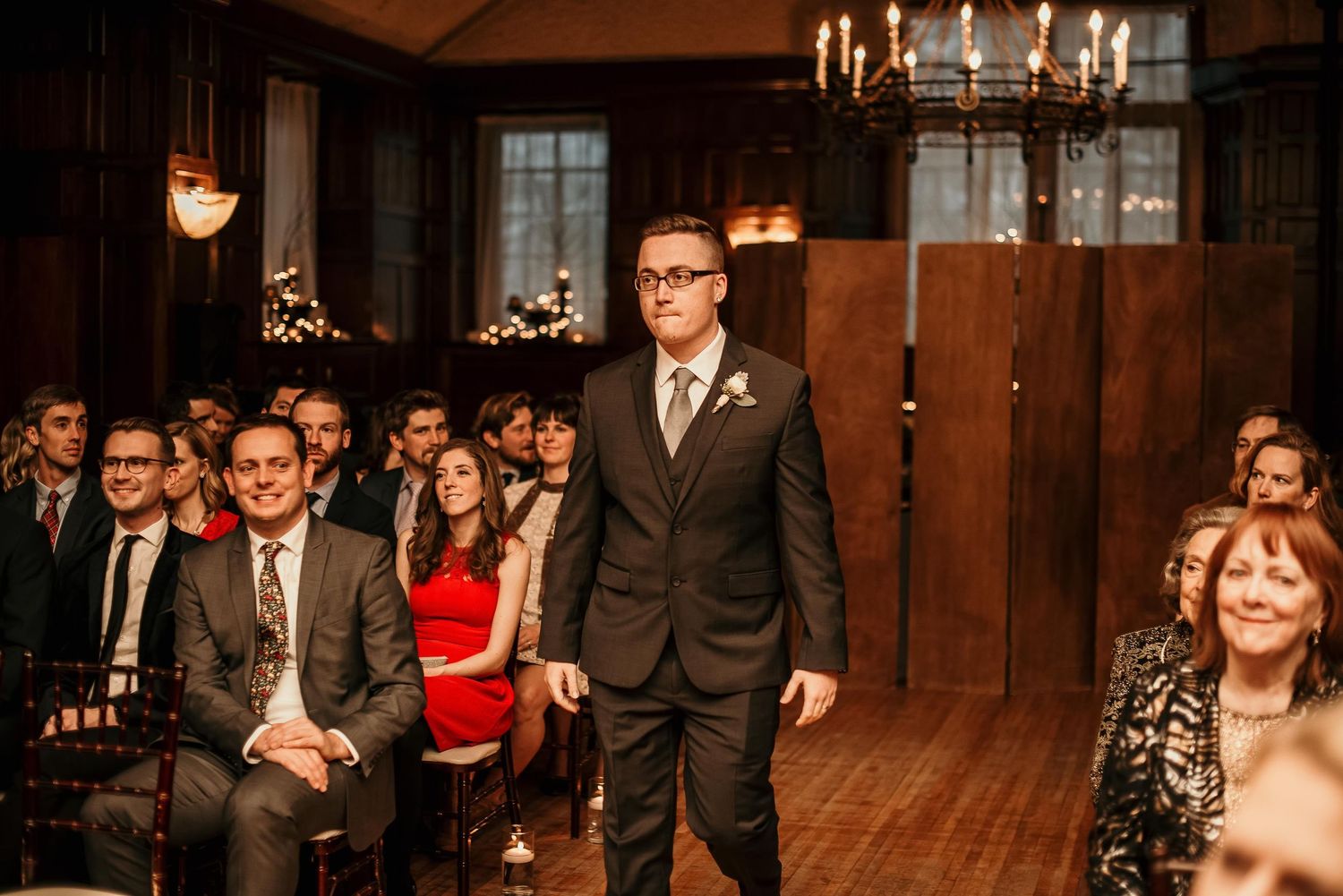 Groom in dark suit walks down aisle at elegant wood-paneled wedding venue with seated guests.