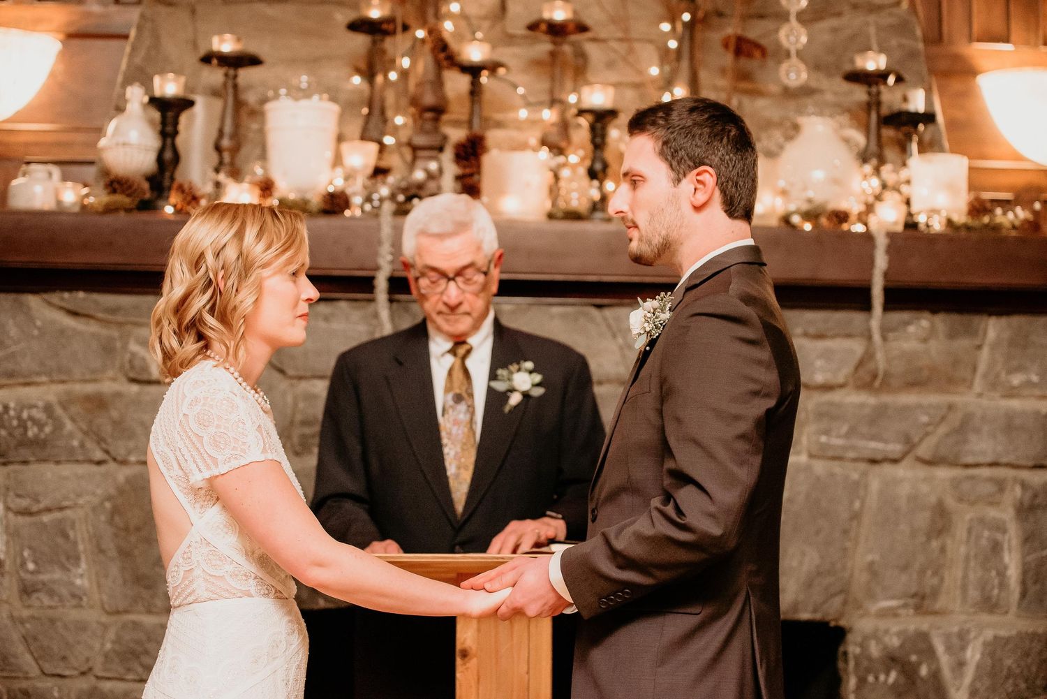 Bride and groom holding hands at rustic stone fireplace wedding ceremony with officiant, warm candlelight ambiance.