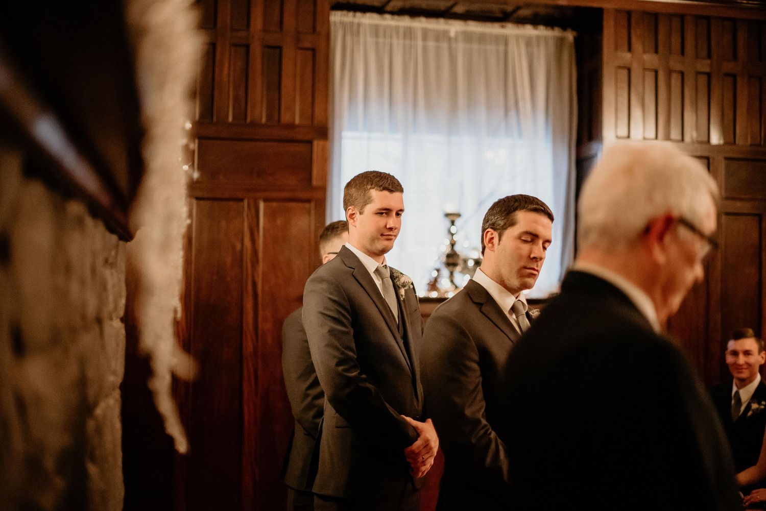 Two groomsmen in gray suits stand at a wedding ceremony inside an elegant wood-paneled room.