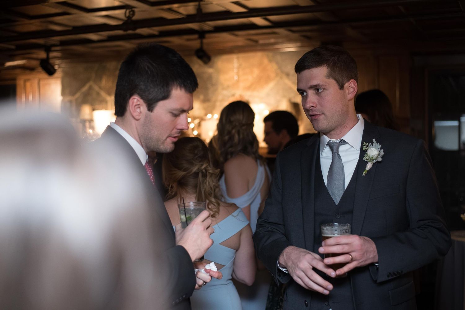 Two groomsmen in suits converse at a wedding reception, one holding a drink, with guests mingling in the background.