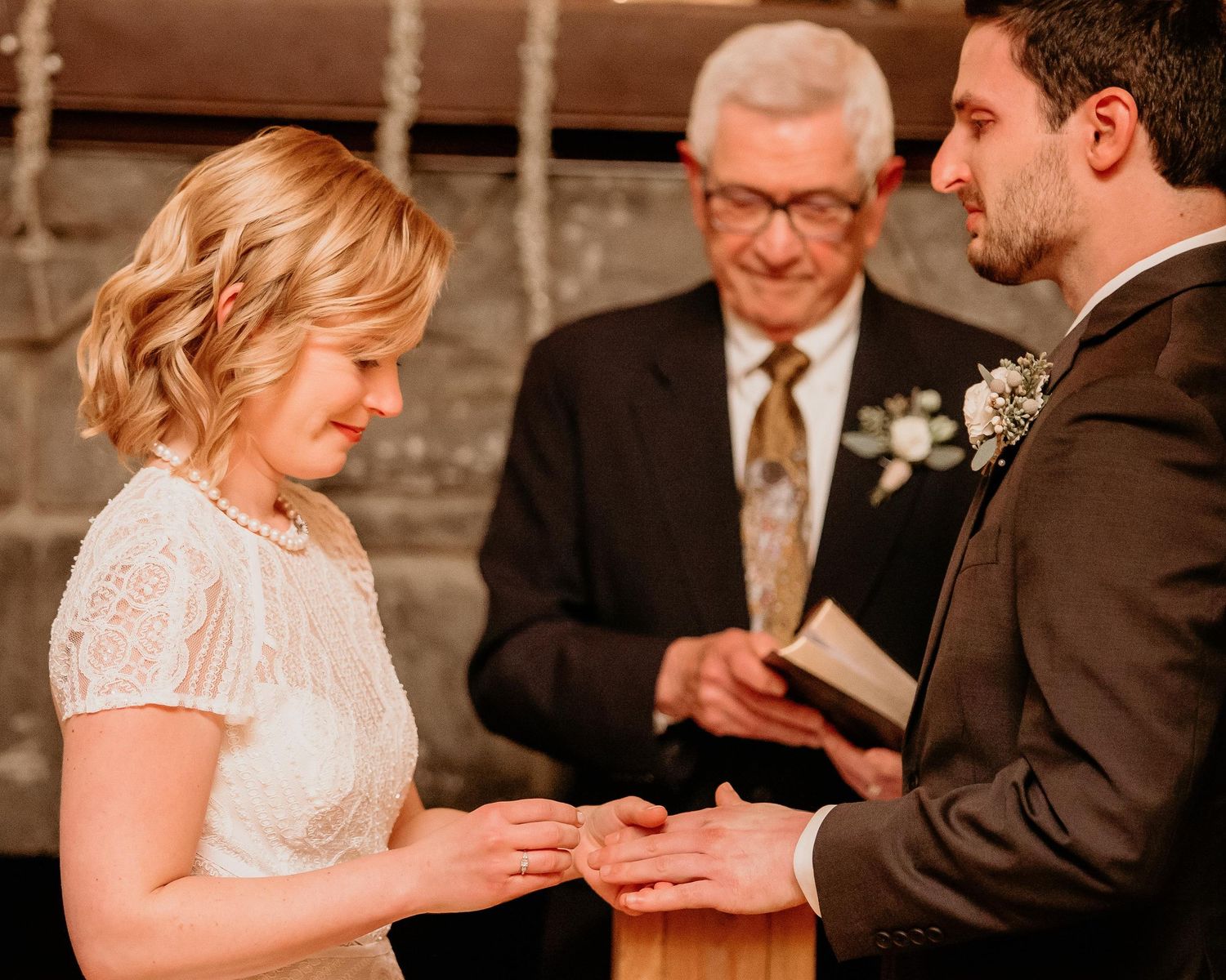 Bride placing wedding ring on groom's finger during ceremony with officiant reading from book in background.
