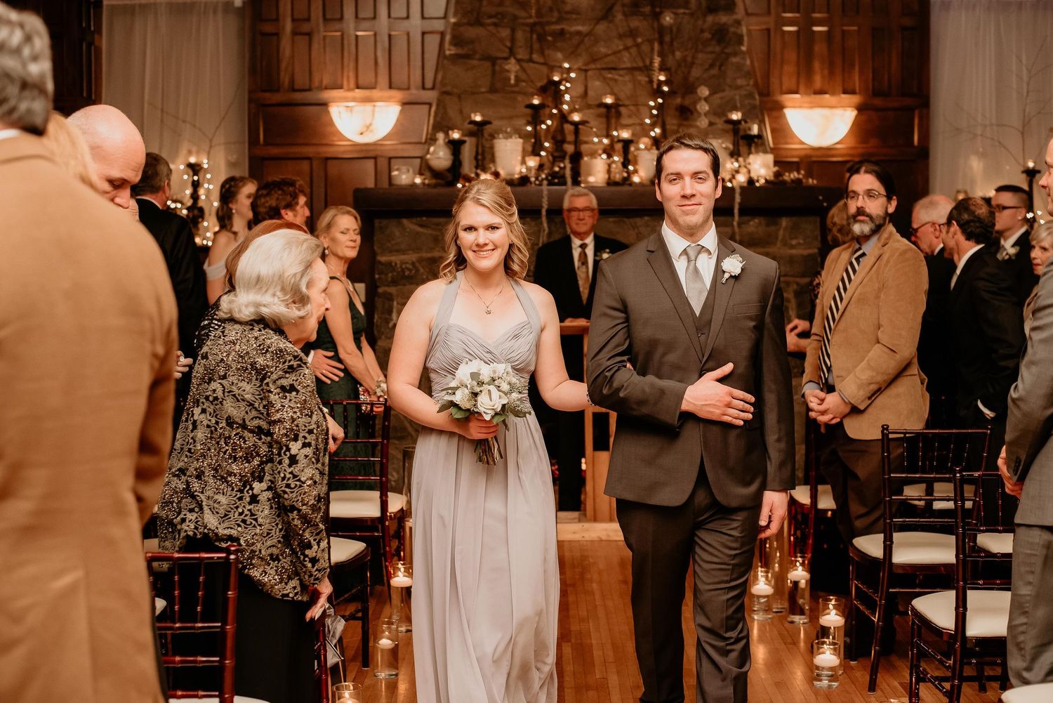A bridesmaid in a gray gown walks down the aisle with a groomsman during an elegant indoor wedding ceremony.