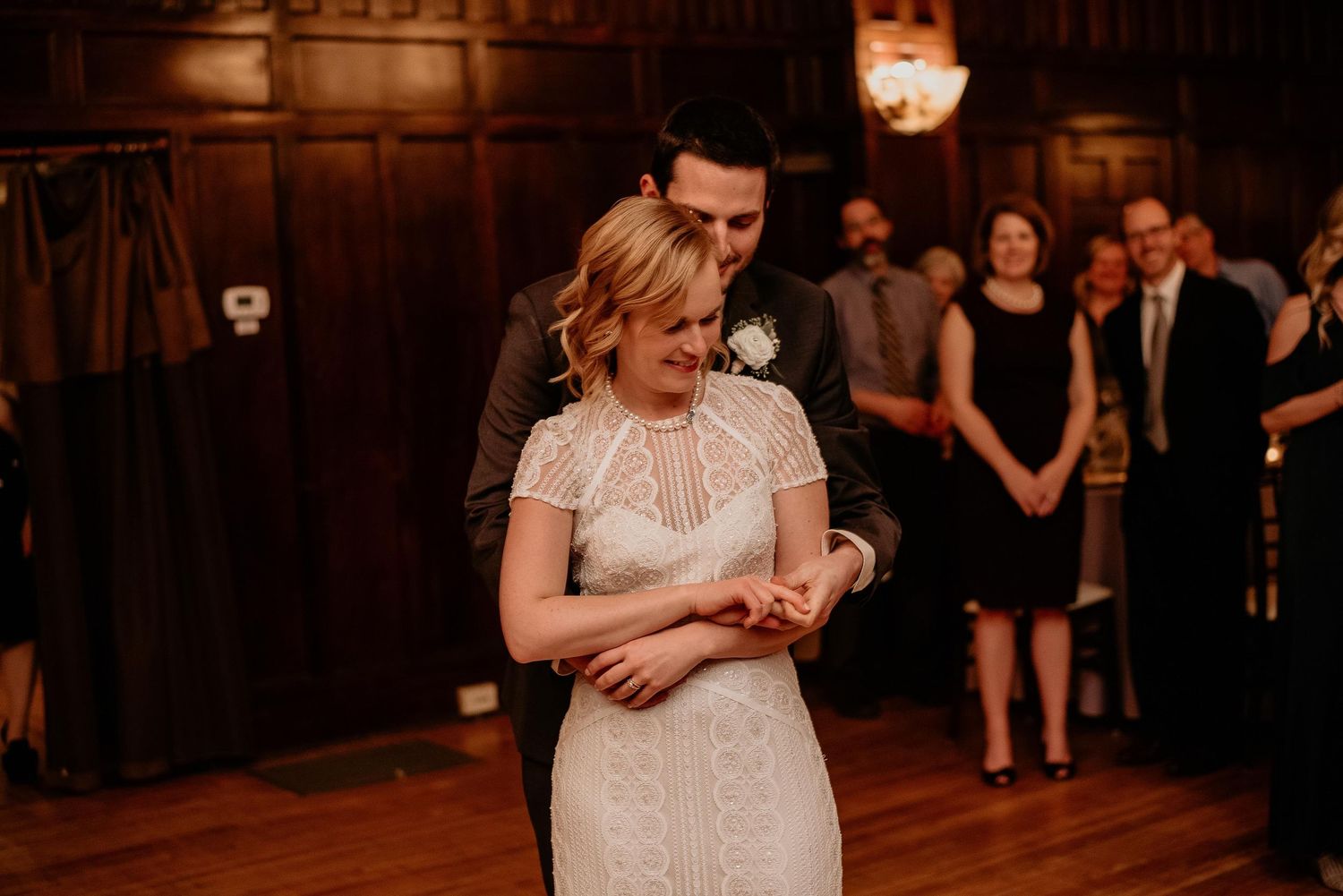 Bride and groom share first dance at wedding reception in elegant wood-paneled ballroom with guests watching.