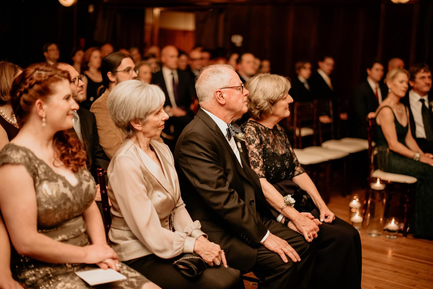 Elegantly dressed wedding guests seated in rows, attentively watching a ceremony in a warmly lit venue.