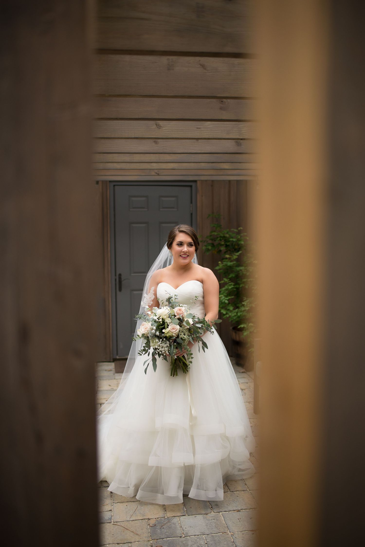 Bride in strapless ballgown wedding dress holding floral bouquet standing before gray door.
