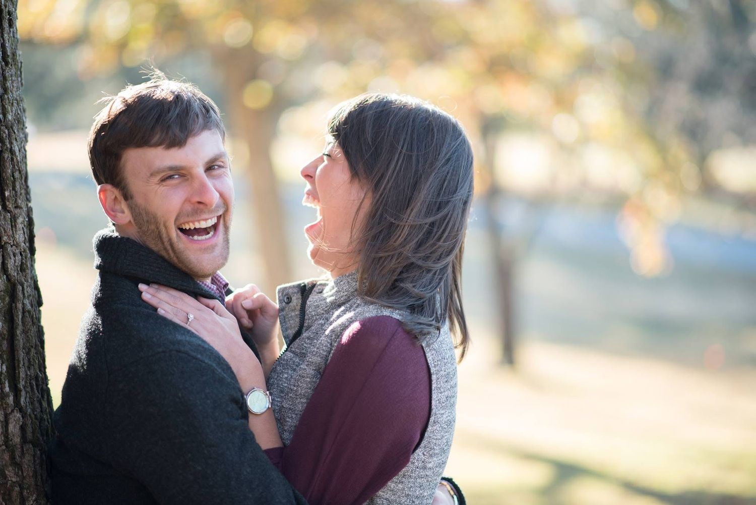 Happy couple laughing together outdoors leaning against a tree in warm autumn sunlight.