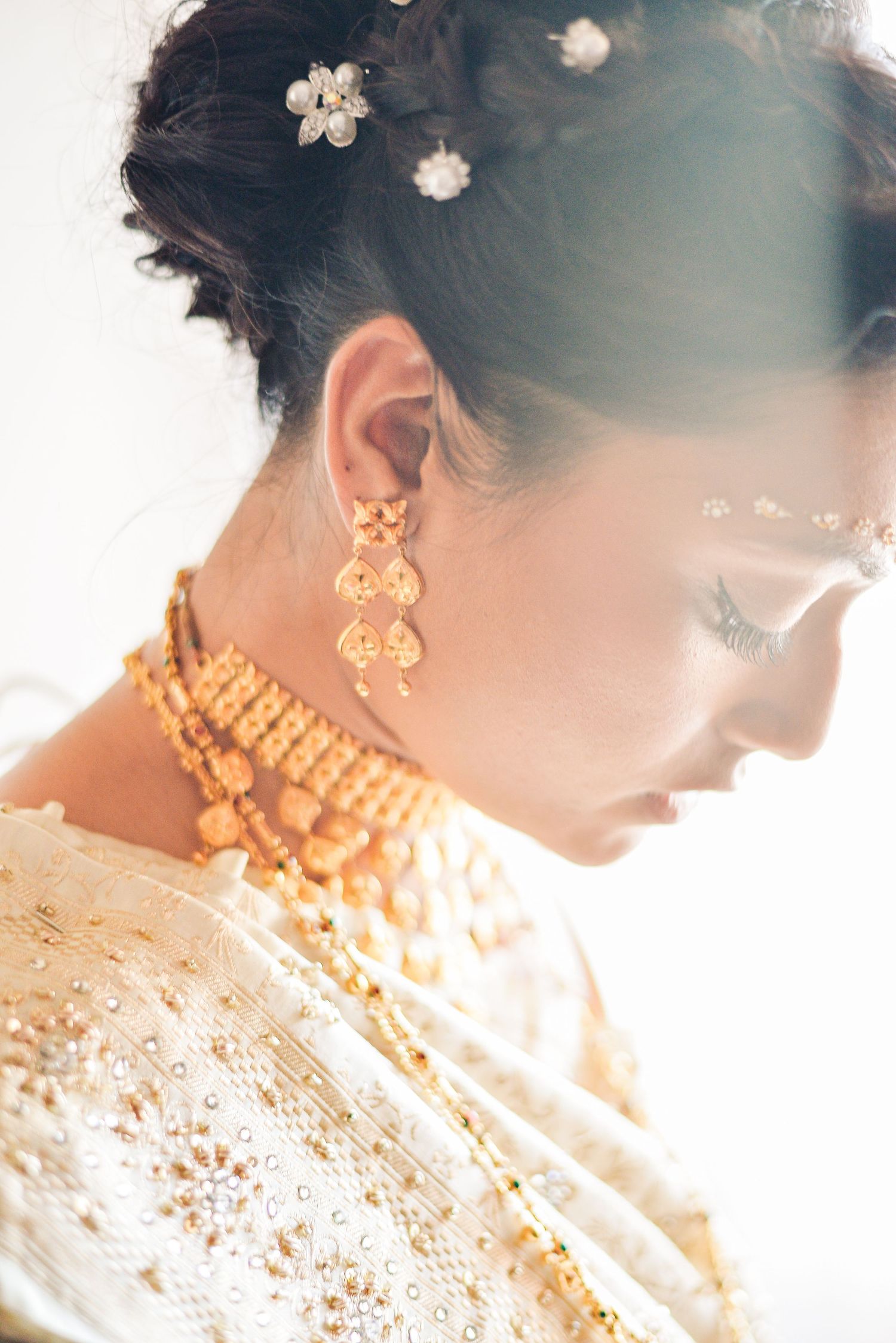 Indian bride in profile wearing ornate gold chandelier earrings and beaded necklace with floral hair accessories.