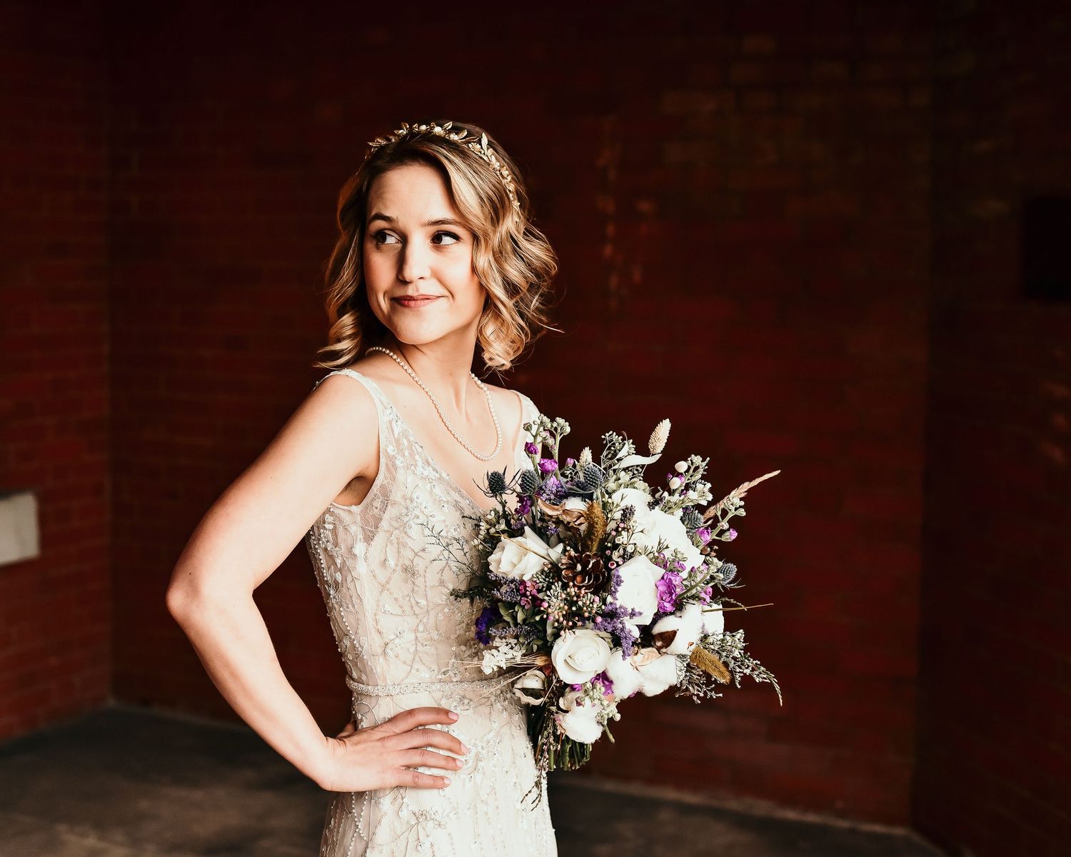 Bride in lace wedding gown holding wild purple and white bouquet against rustic brick wall backdrop.