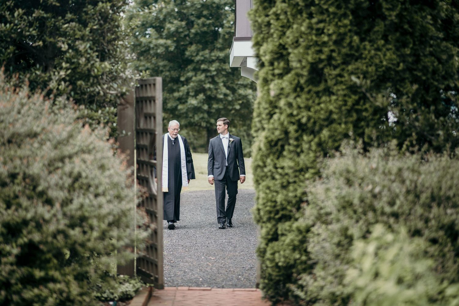 Two men in suits walking through garden pathway lined with hedges.