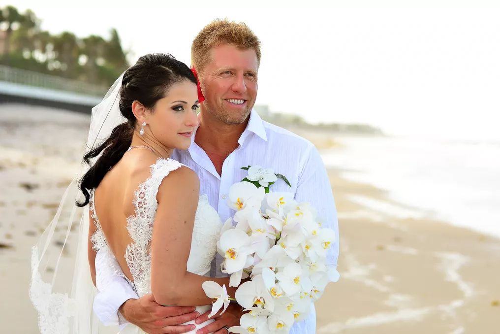 Bride and groom posing on a beach wedding with white orchid bouquet and ocean backdrop.