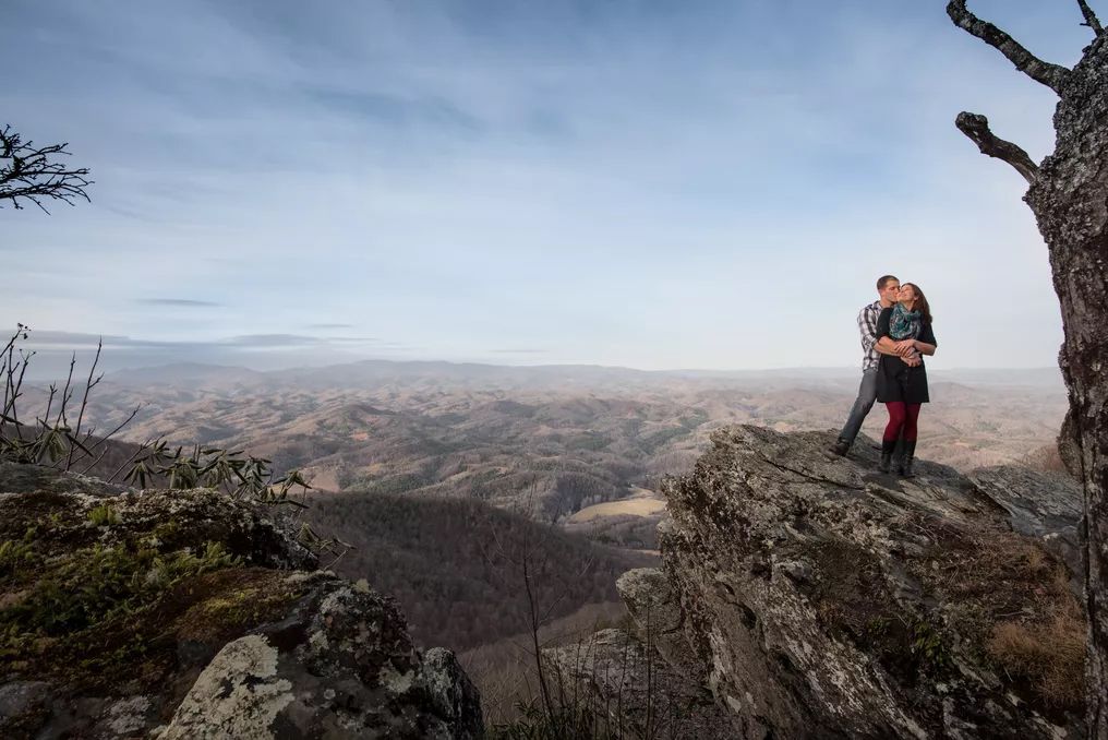 Couple shares a romantic kiss on a rocky mountain summit with a breathtaking panoramic landscape behind them.