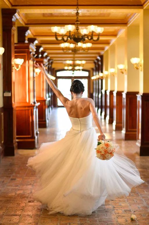 Bride in white gown walks down elegant hallway, holding bouquet, one arm raised, in a grand venue with chandeliers.