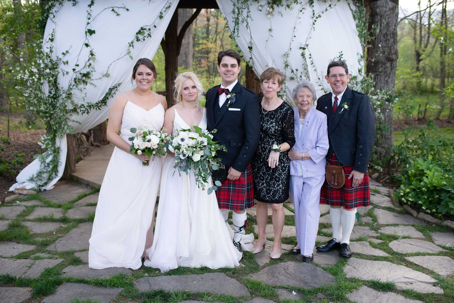 Wedding party posing outdoors, bride and groom in kilt with family members in formal attire near floral arch.
