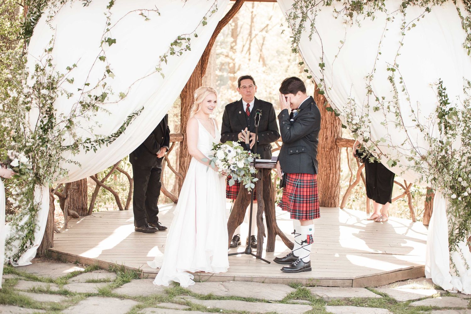 Bride and groom exchange vows under floral arch with officiant, groom wearing tartan kilt at outdoor wedding ceremony.