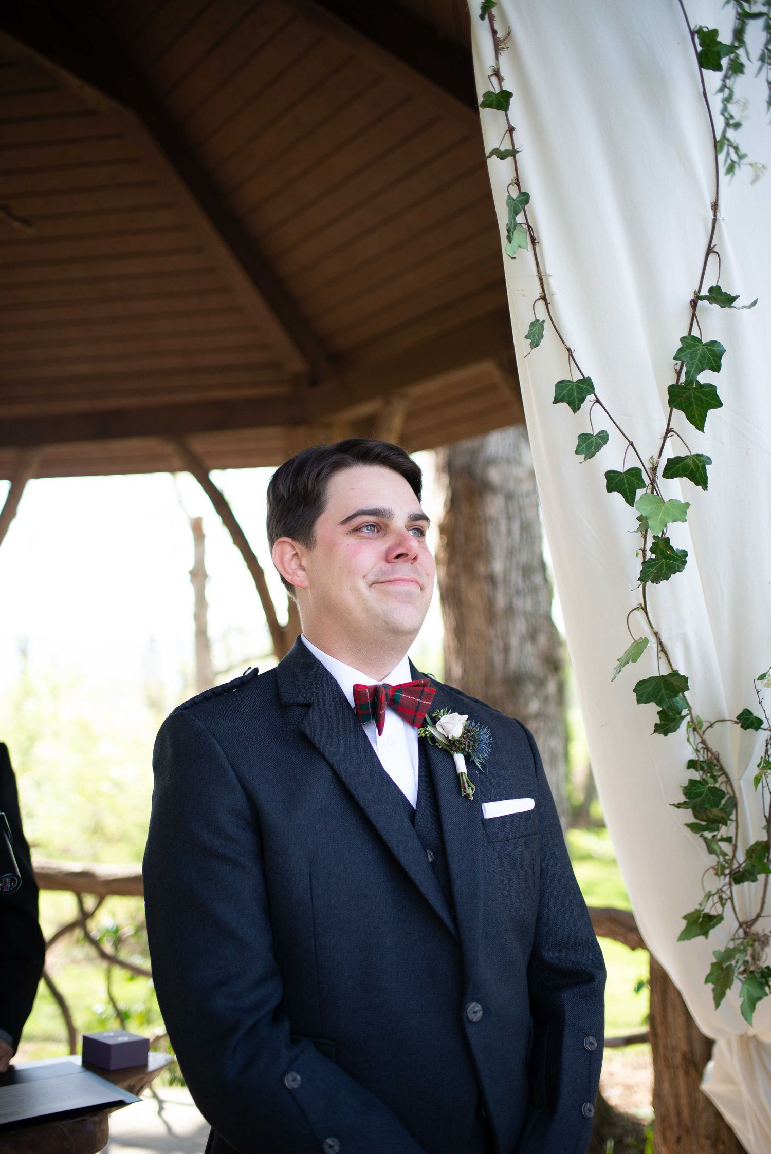 Groom in navy suit and plaid bow tie smiling emotionally at wedding altar decorated with ivy and white draping.