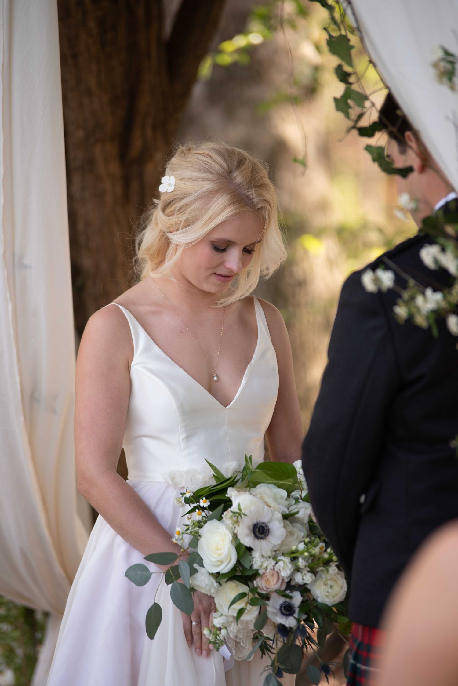 Bride in white gown holding white floral bouquet with anemones and roses during outdoor wedding ceremony.
