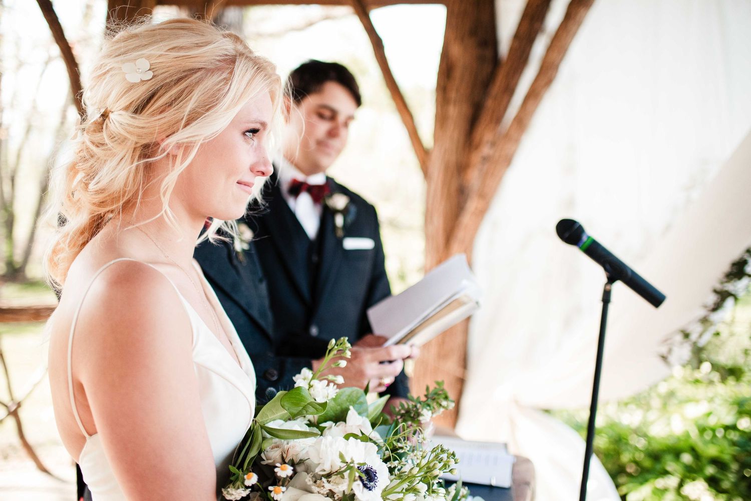 Bride holding bouquet reads vows at outdoor wedding ceremony while groom stands behind her at microphone.