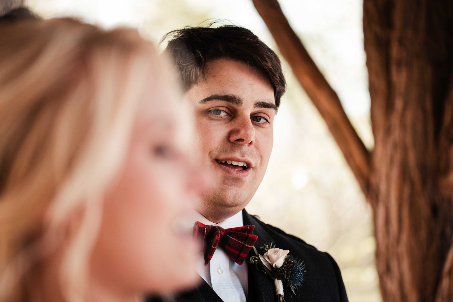 Groom in black tuxedo with red bow tie gazing lovingly during outdoor wedding ceremony.
