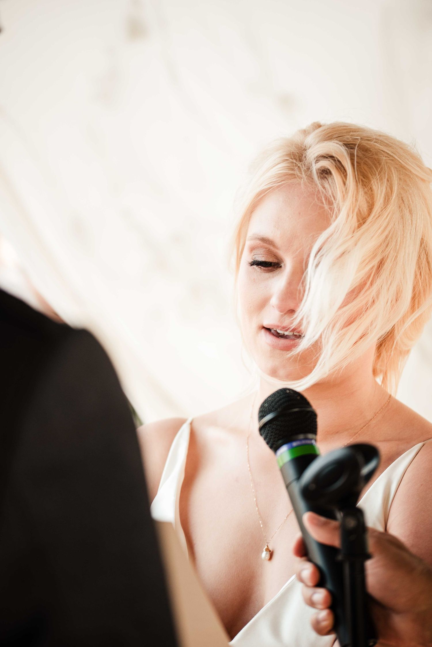 Blonde woman speaking into a black microphone at an indoor event, wearing a white dress.