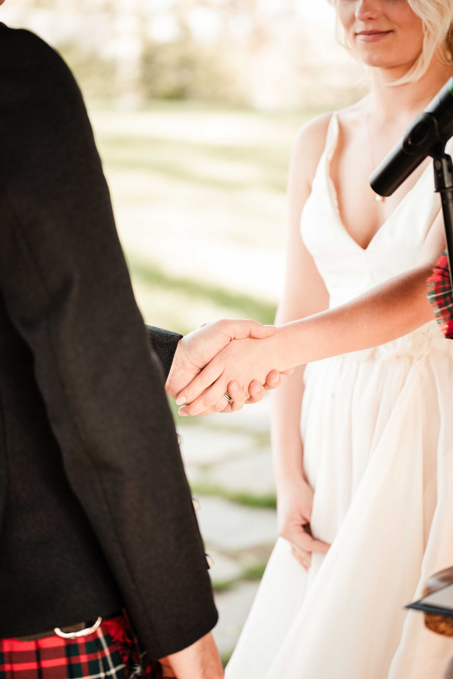 Bride in white dress and groom in tartan kilt holding hands during outdoor wedding ceremony.