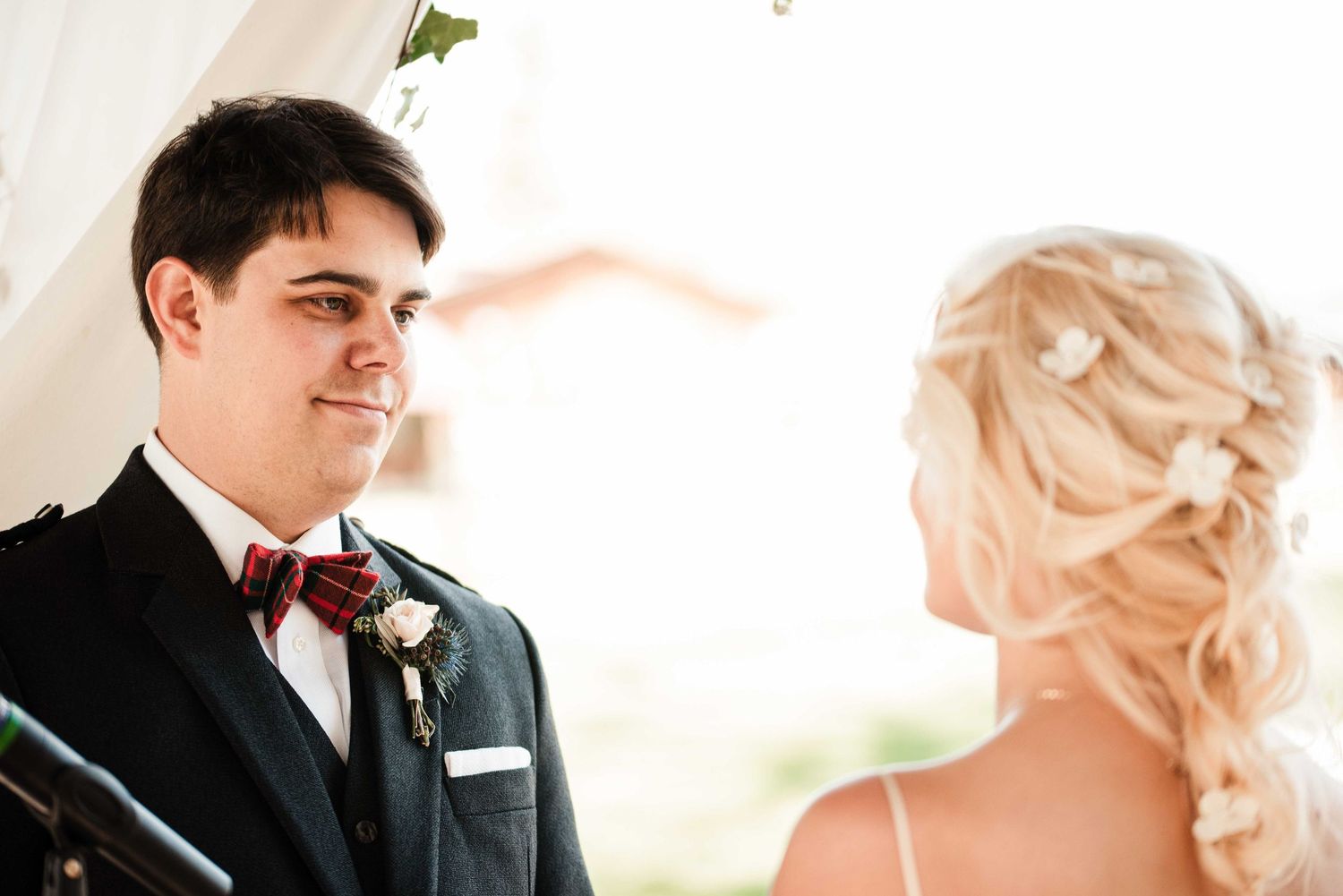 Groom in dark suit with burgundy bow tie smiling at blonde bride during outdoor wedding ceremony.