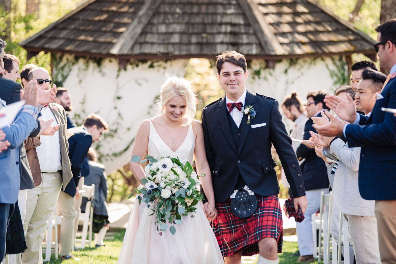 Bride and groom walk down the aisle, groom wearing a red tartan kilt, guests celebrating outdoors under a floral gazebo.
